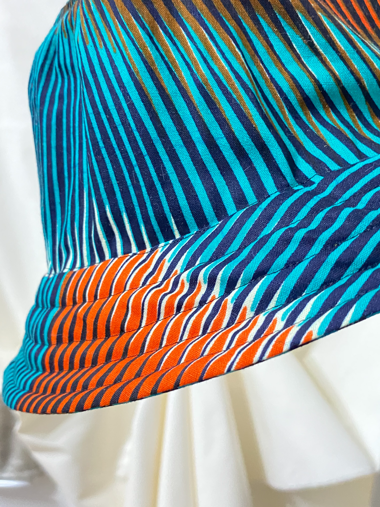 Close up of bucket hat named Bonaberi. Fabric is a turqouise tint with wave like lines in a darker turqouise and hues of orange.